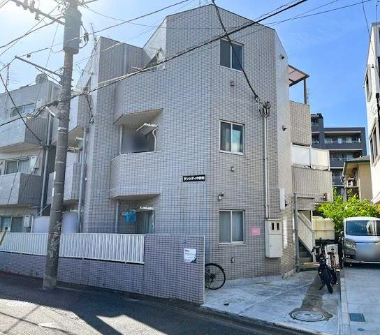 白いタイル張りの三階建ての建物と、前に自転車とバイク、駐車している車がある。周囲に電線や電柱が見える都市の風景。
