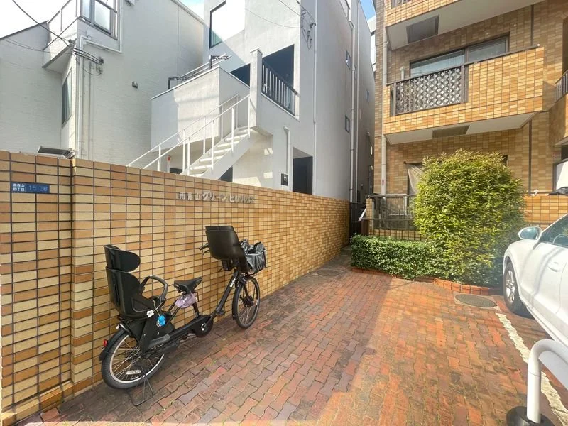ブロック積みの壁の前に自転車と車が parkedしている、マンションの駐車場の風景。背景には建物と植物が見える。