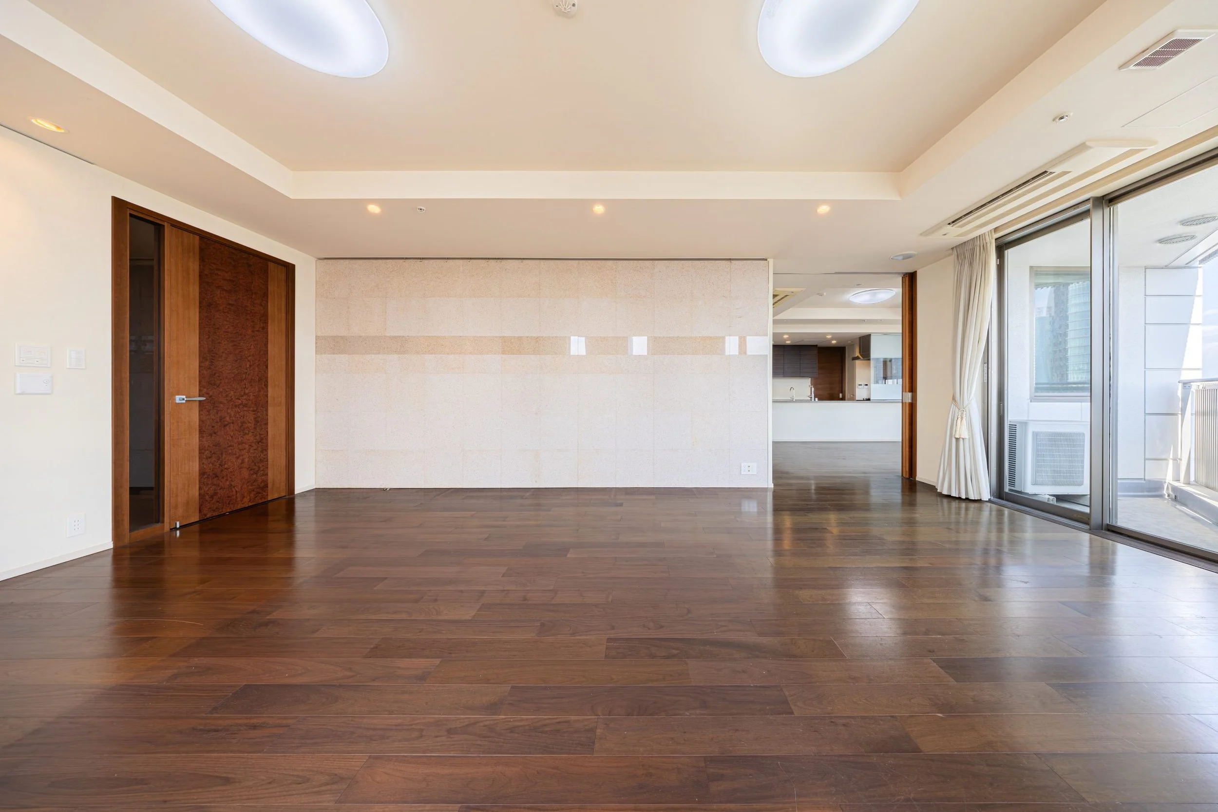 空のリビングルーム。木製のフローリングと白い壁、スライドドアと大きな窓から自然光が入っている。リビングルームとキッチンエリアが見える。