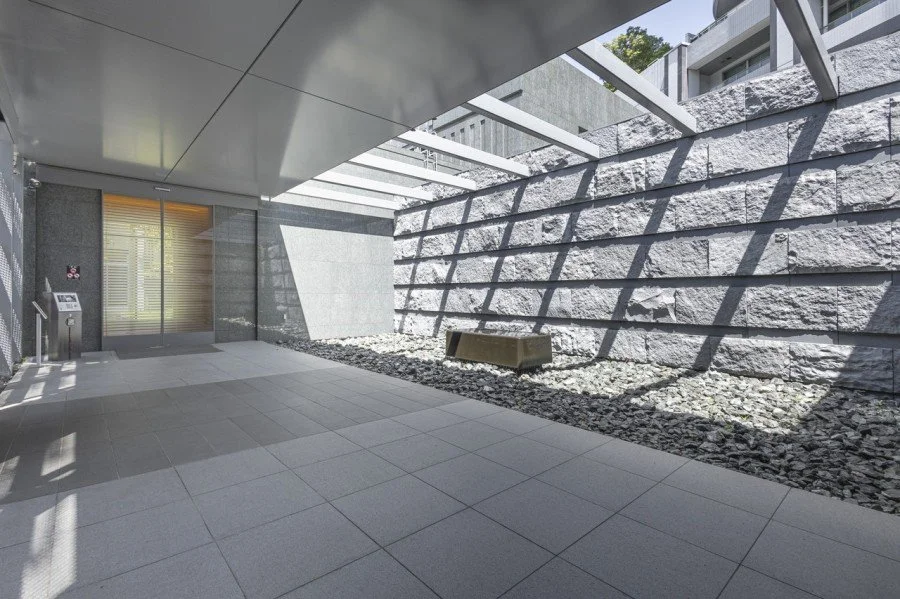 建物の出入口付近の広い屋外空間。左側にガラス扉とインターホン、右側に大きな石の壁と小さな砂利が敷かれたスペースがある。空中からの光と影が交差している。