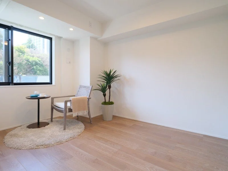 白い壁と木の床の部屋に、グリーンの観葉植物と小さなテーブルと椅子がある。窓から外の緑が見える。