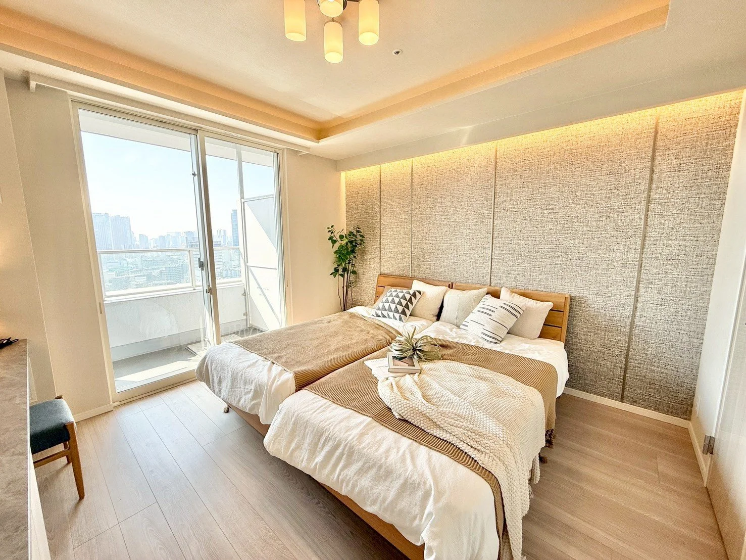 明るい光の差し込む洋室のベッドルーム。大きな窓から都市の景色が見える。ベッドには複数の枕とカバーが掛かっている。室内には観葉植物も置かれている。