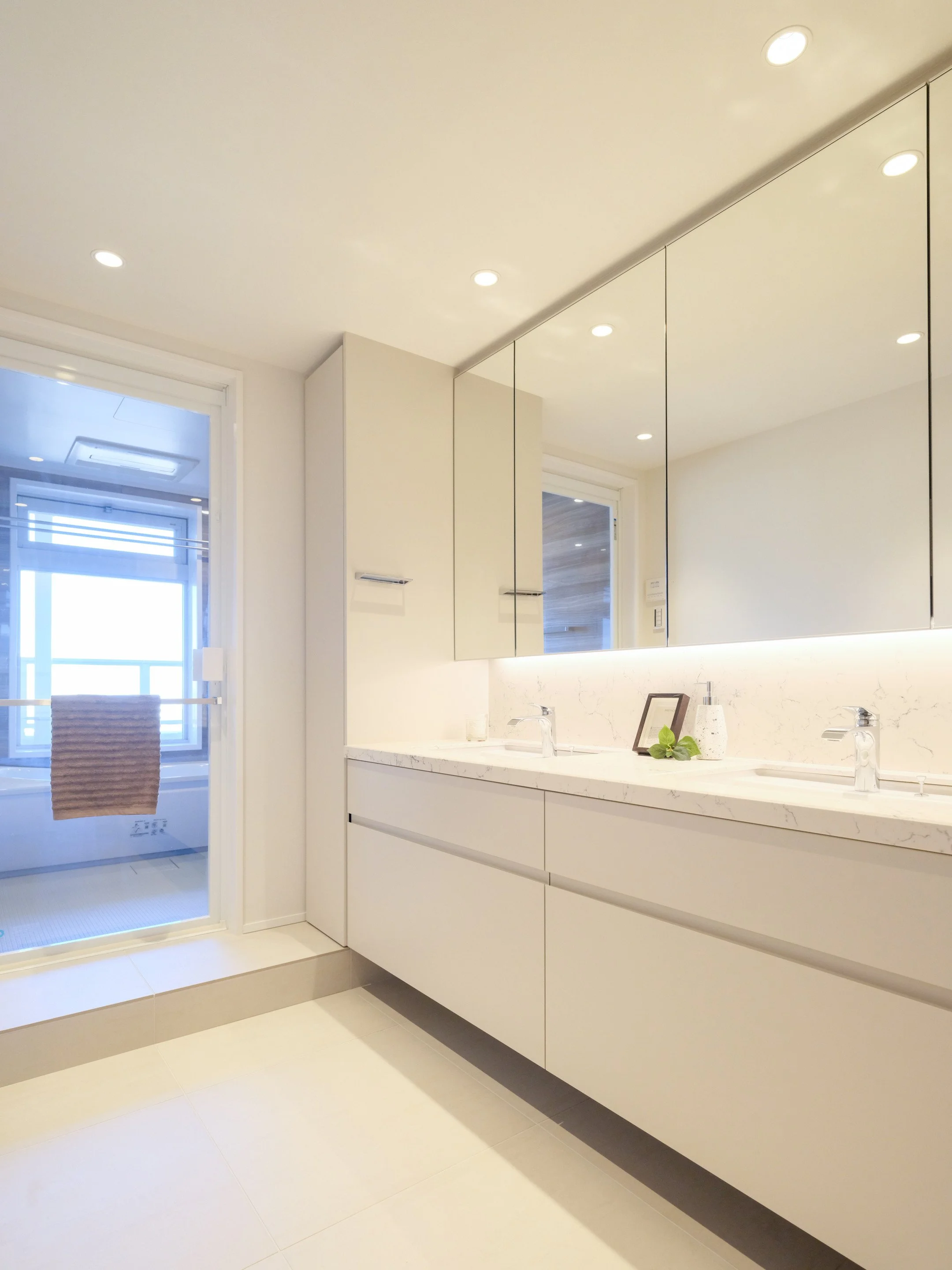 白を基調とした洗面台のあるシンプルなバスルーム。鏡張りの収納棚と二つの洗面台、窓からの自然光が差し込む