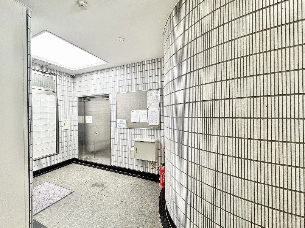 白いタイル張りの壁と金属製のエレベーター扉があるエレベーターホール。左側に窓と掲示板があり、下に消火器が置いてある。天井には照明がついている。