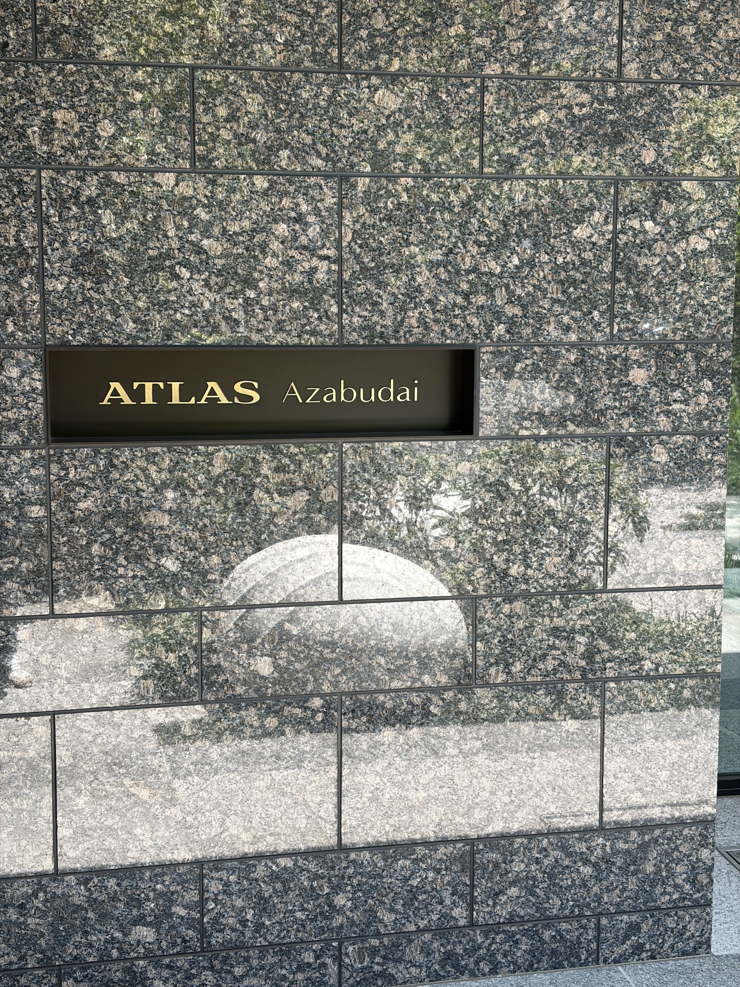 黒い背景に金色の文字で「ATLAS Azabudai」と書かれた看板が、 granite タイルの壁に取り付けられている。