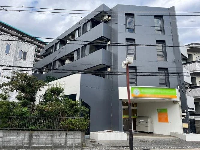 灰色の三階建ての建物。下部にはコンビニエンスストアの看板と自動販売機があり、周囲には木々とフェンスがある。