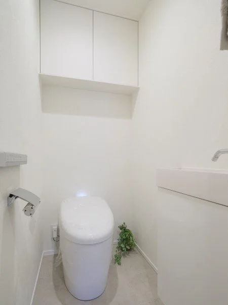 白い壁のトイレルーム。トイレットペーパーと緑の植物、上部に収納棚があるシンプルな空間です。