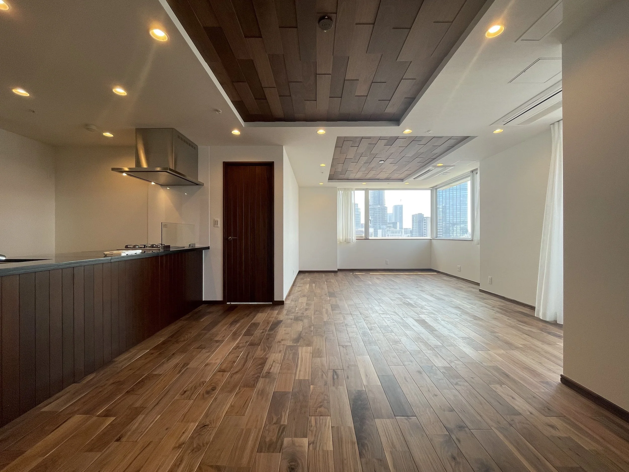 高層ビルの景色が見える大きな窓のある空っぽのリビングルーム。木製の床と天井のデザインが特徴的で、明るい自然光が入っています。