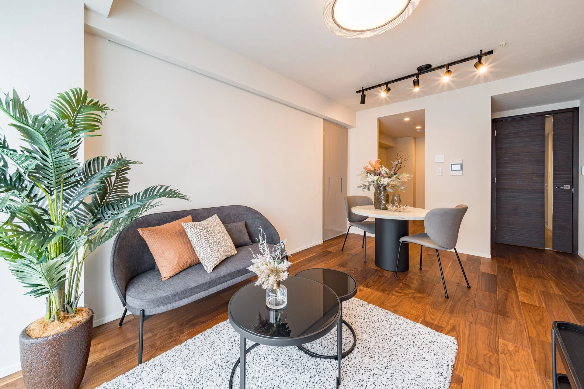 モダンなリビングルーム、グレーのソファと多色のクッション、丸いコーヒーテーブルとラグ、ダイニングエリアに花のデコレーションと椅子、木製のフロアと暗いドア