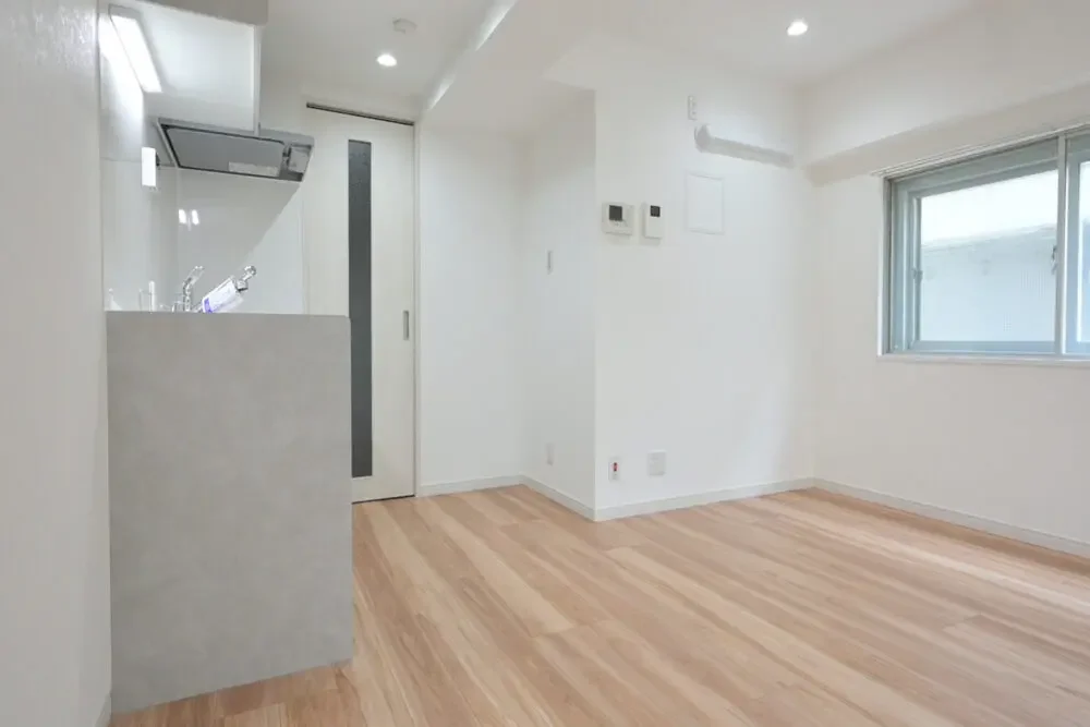 白い壁と木製の床の空間に窓があり、キッチンとドアが見える部屋