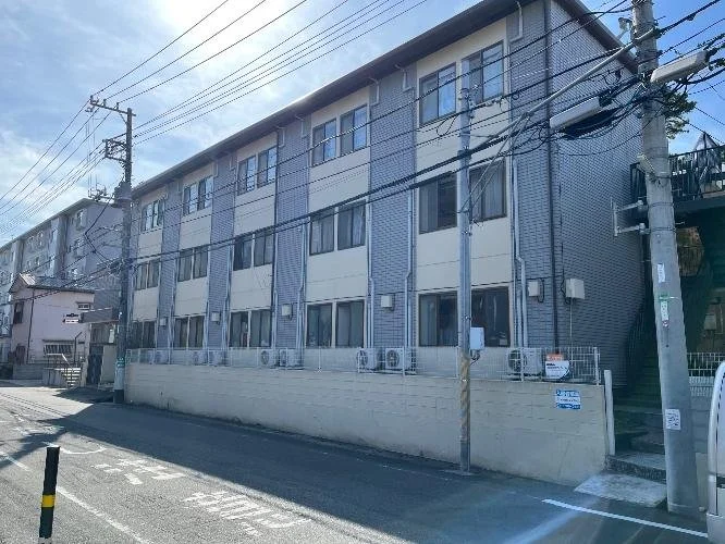 三階建てのアパートまたはマンションの外観。道路側から見た灰色と白の建物で、多くの窓と空調ユニットが設置されている。