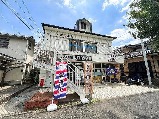 二階建ての建物とその前の駐車場。建物には「大野ビル2」と書かれ、下階には美容院とサロンの看板。階段は外側にあり、旗には「営業中」と書かれている。