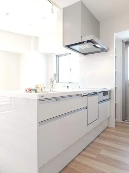 白いキッチンカウンターと収納、シルバーの換気扇と窓、木製の床の現代的なキッチン室内