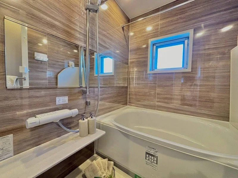 木目柄のタイル壁の浴室で、白い浴槽と鏡と小さな窓があります。