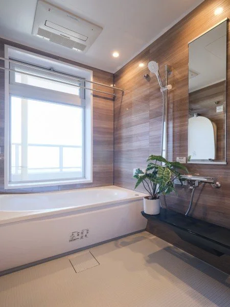 木目調の壁の浴室、白い浴槽、窓、シャワー、鏡、バスルームに置かれた植物。