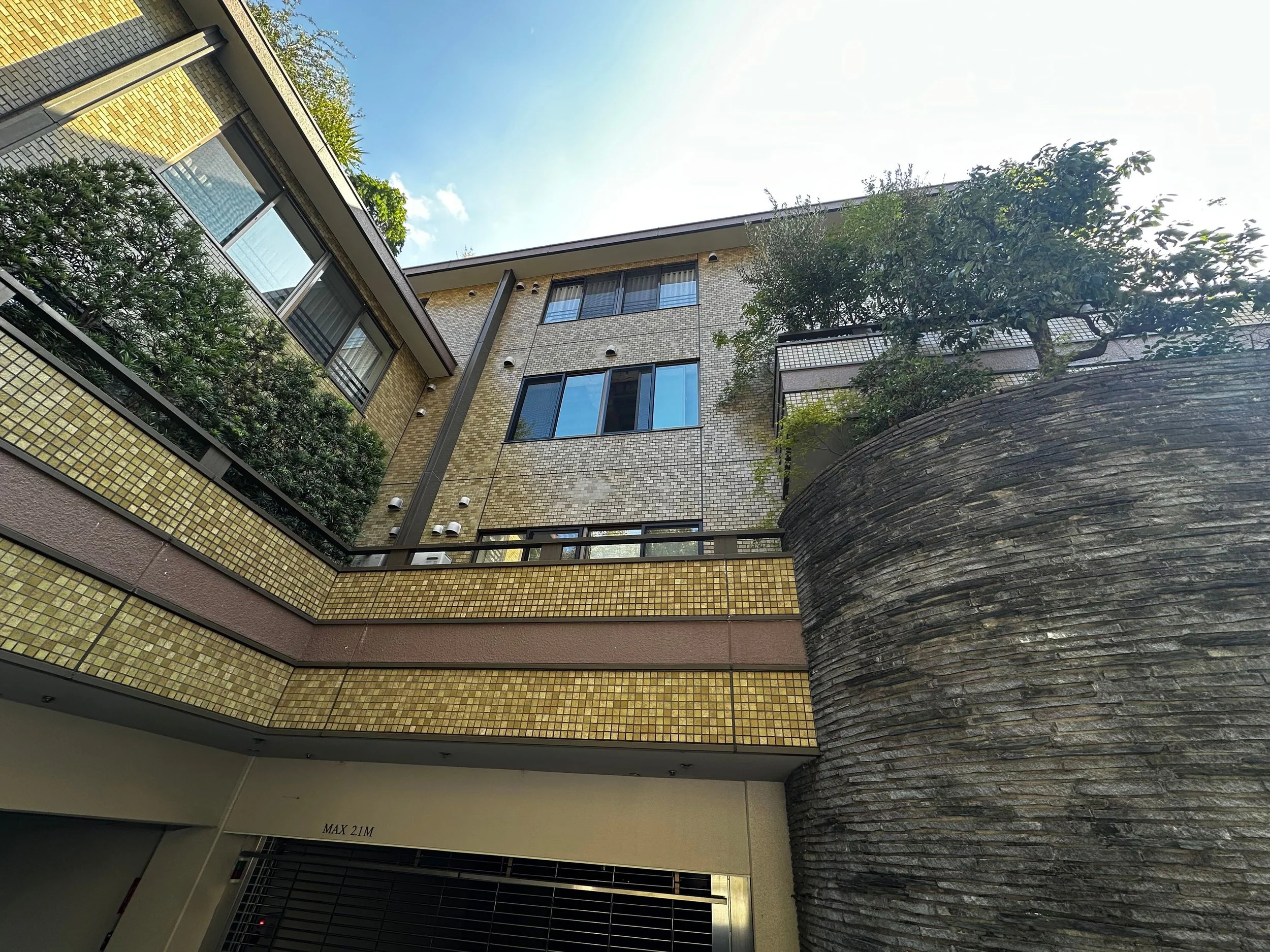 複数階建ての建物の外観。レンガとタイルの壁、窓、緑の樹木と青空が見える。
