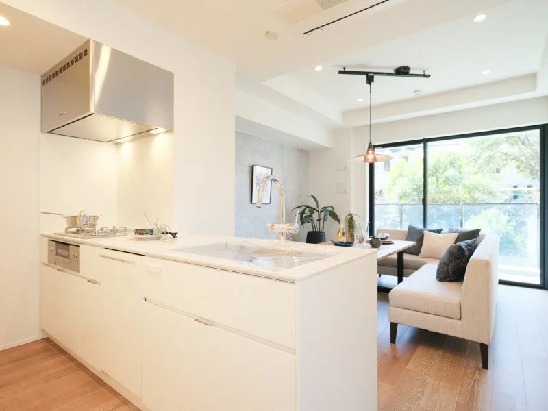 シンプルでモダンなリビング兼キッチンスペース。白いカウンターキッチンとリビングにはソファとテーブル、窓からは緑の景色が見える。