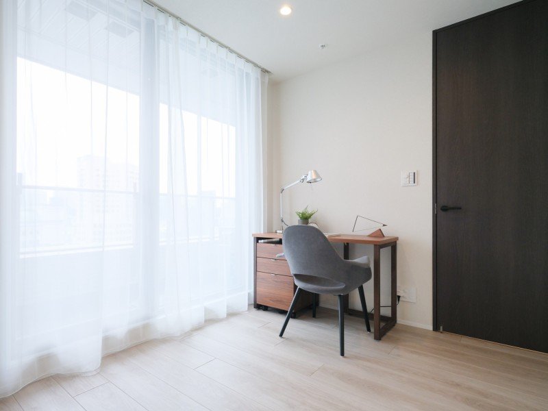 自然光が差し込むシンプルなワークスペース。デスクと椅子、書類ラック、テーブルランプ、観葉植物。白いカーテンと木目調の床と家具が調和した静かな部屋です。