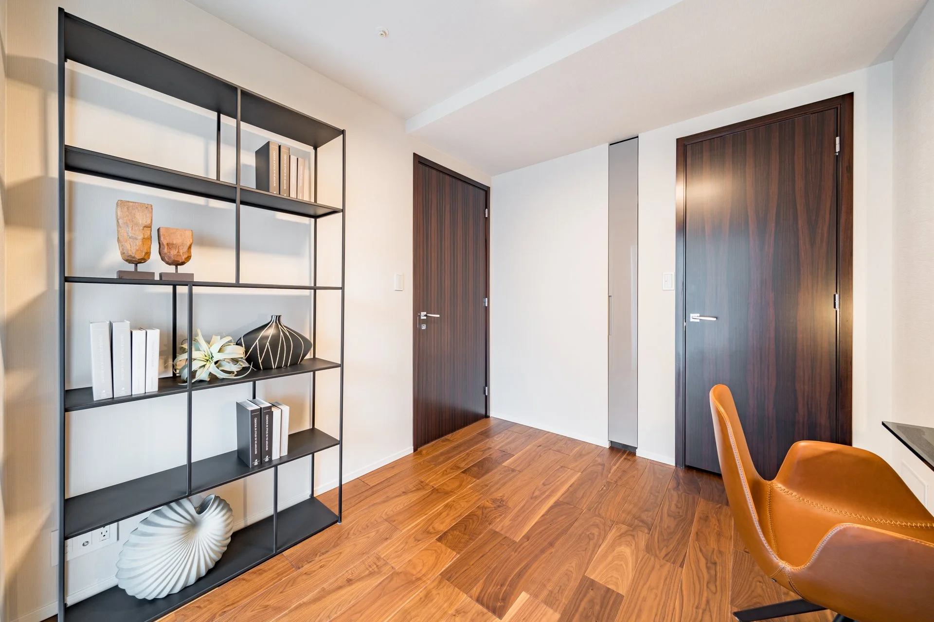 木製の床と白い壁の部屋に、黒いスチールの本棚と茶色の革張り椅子が置いてある風景