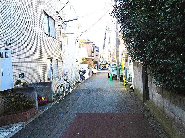 狭い住宅街の道路、小さな車や自転車、コーンが並び、住宅の壁や電線が見える風景