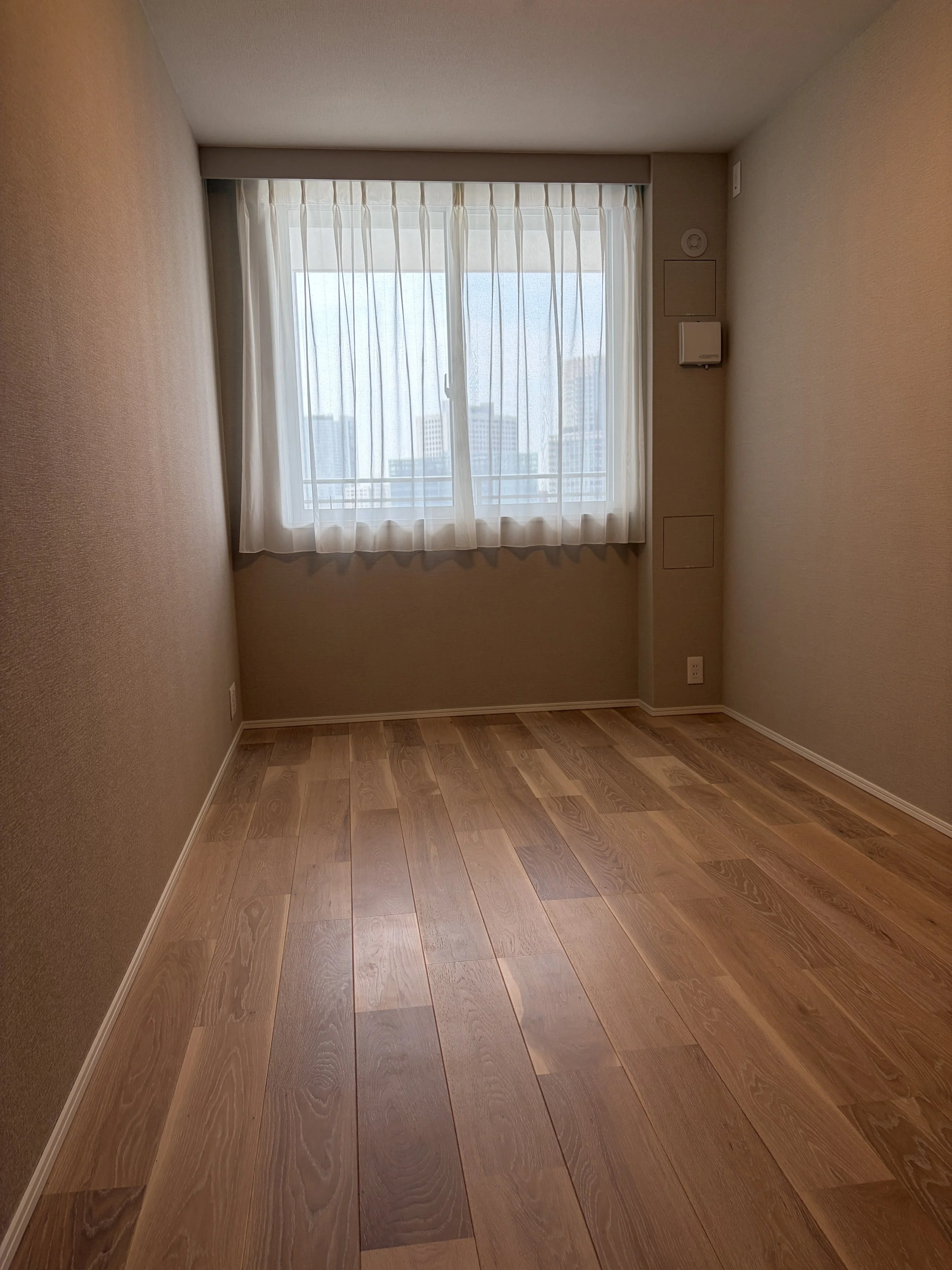 木製の床の部屋に白いカーテンを通した窓があり、外のビル群が見える。壁には電気コンセントと換気扇が設置されている。