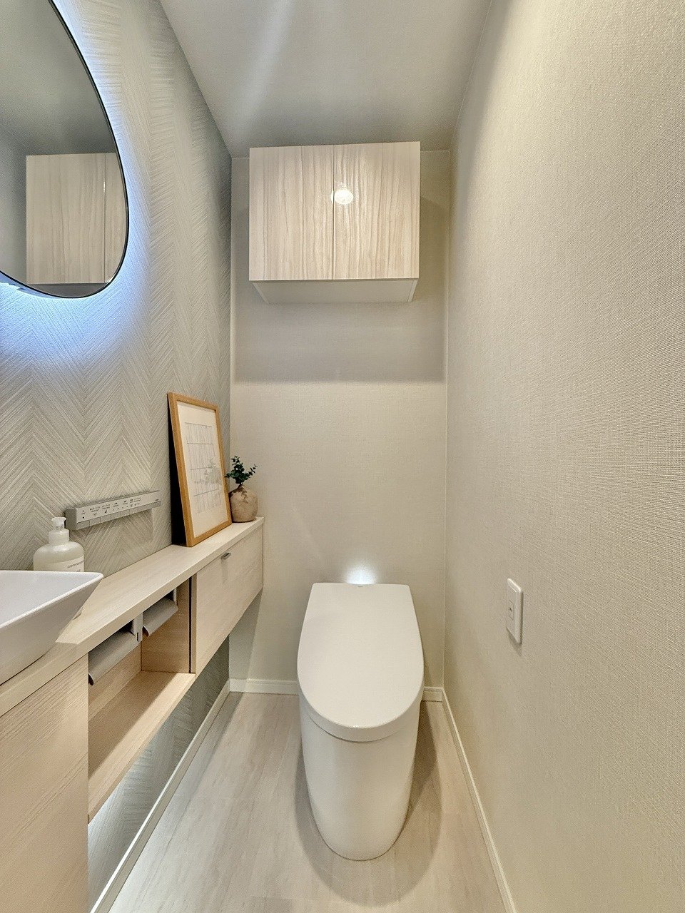 シンプルでモダンなトイレの空間。白い便器と木製の収納棚、小さな飾りと置物が置かれている。鏡と手洗い用の液体石鹸も見える。