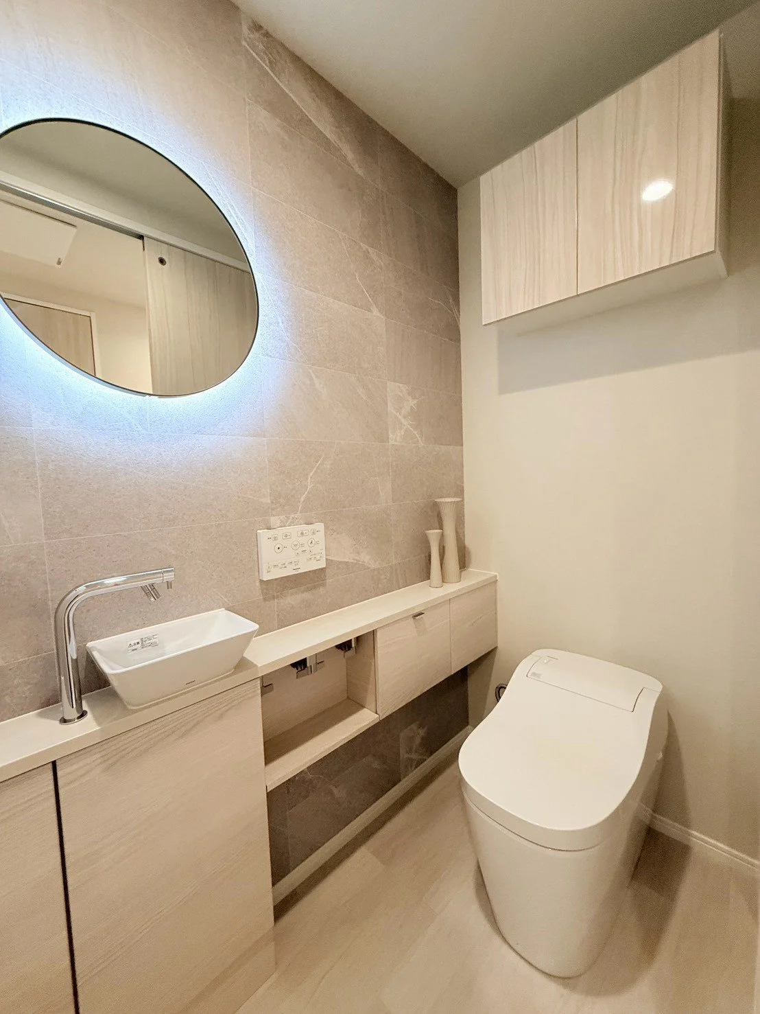 モダンな洗面所とトイレの空間。丸い鏡、シンプルな洗面台、トイレ、収納棚が配置されている。