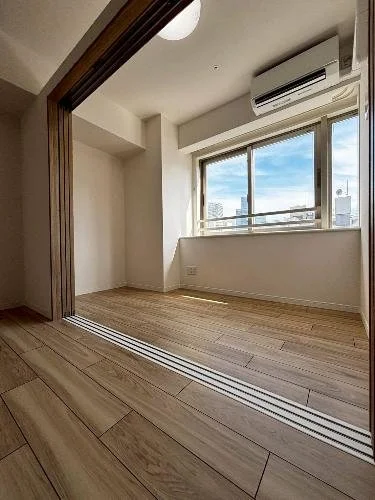 窓から都市の景色が見える空室の部屋です。エアコンと木製の床があります。