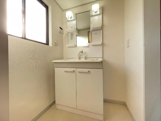 白い洗面台と鏡のあるシンプルな洗面所。窓と照明あり。