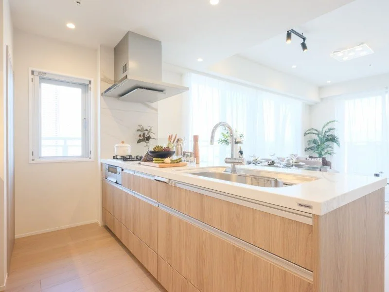 明るいキッチンで、窓から自然光が入り、木製のキャビネット、シンク、コンロ、調理器具、植物の飾りがある。