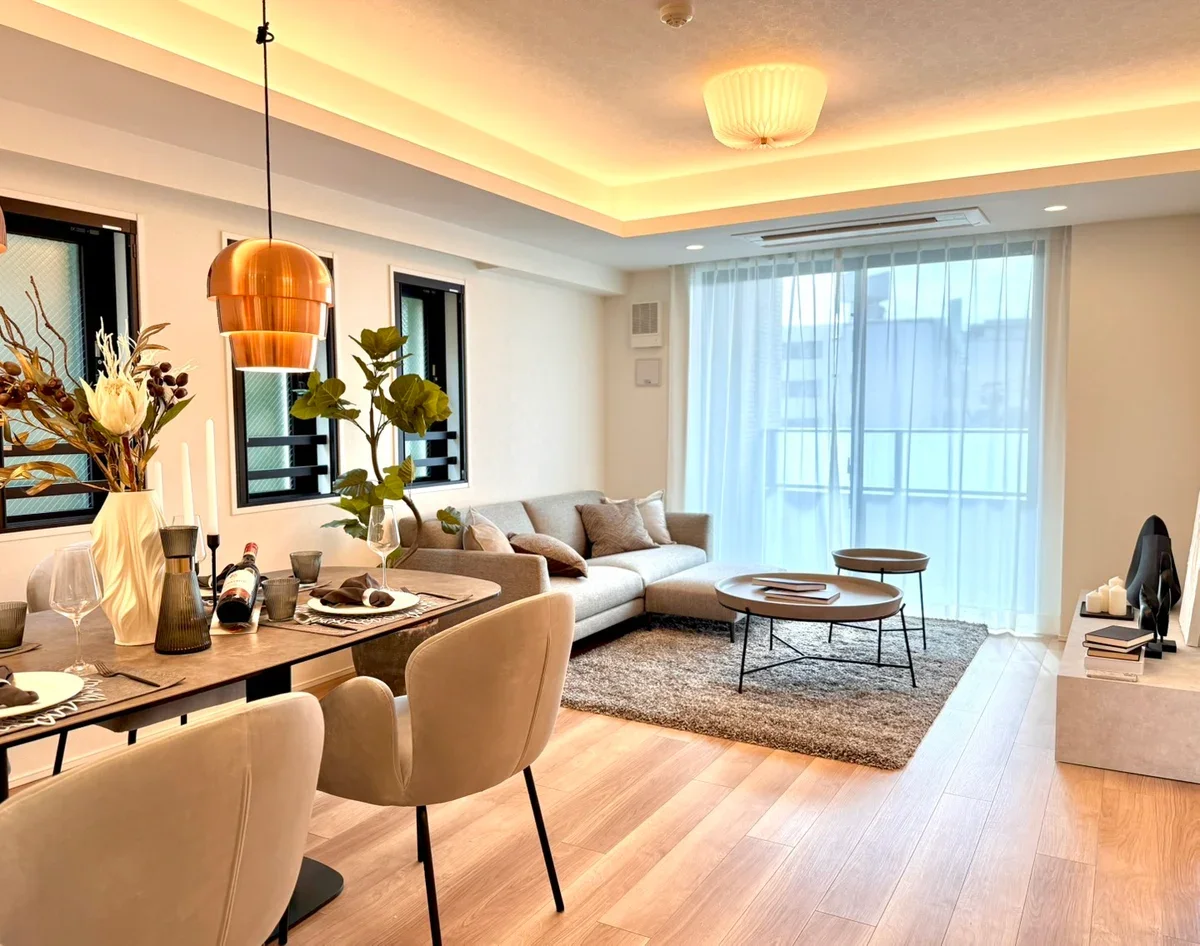 リビングルームに大きな窓とカーテン、ソファ、コーヒーテーブル、床にラグ、小さな丸いサイドテーブル、ダイニングテーブルと椅子、花瓶に花とキャンドル、壁にアート作品、ブラウンの照明、木製の床