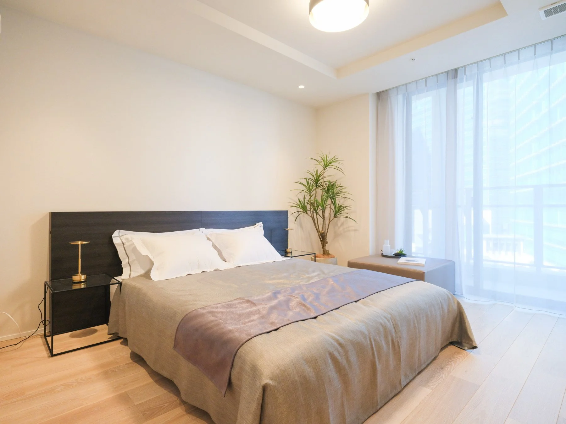 明るい自然光が差し込むシンプルでモダンな寝室。ダークウッドのヘッドボードと白い寝具。サイドテーブルには小さな金色のランプ。観葉植物と窓辺のベンチに置かれた小物と本。薄いカーテンが外の景色をやわらかく覆っている。