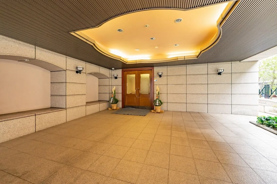 エントランスの入口、木製のドアと二つの縦長の飾り花、壁に取り付けられた照明と天井の照明がある屋外のポーチ部分