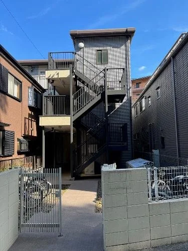 中規模の鉄製の外階段がついた三階建ての住宅。周囲はコンクリートの塀と小さな門とフェンスに囲まれている。