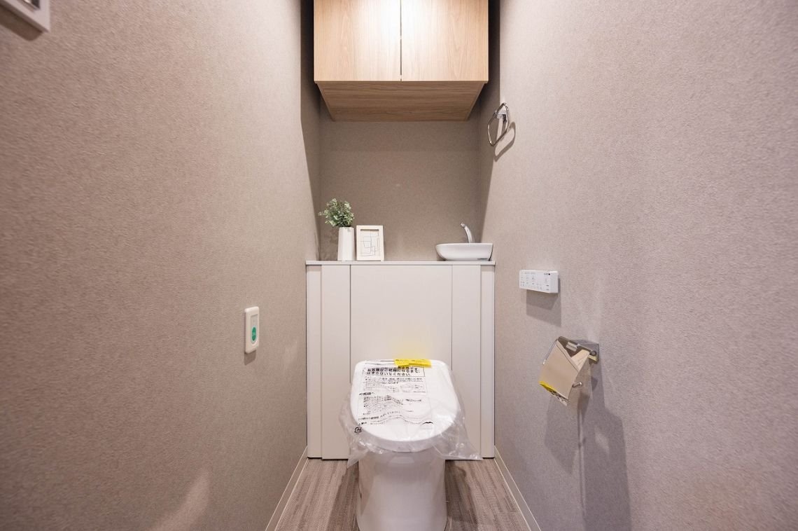 トイレの内部で、便座と小さな洗面台、棚、壁掛けの紙巻き器とリモコンが見える。