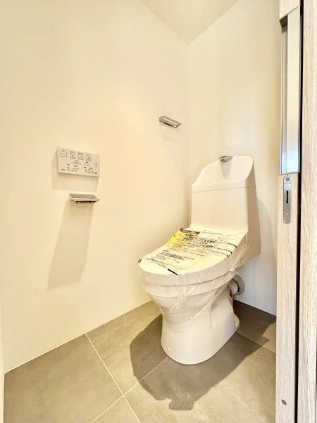 白い壁と便座のトイレの部屋。トイレットペーパーのホルダーとリモコンのようなものが壁に取り付けられている。