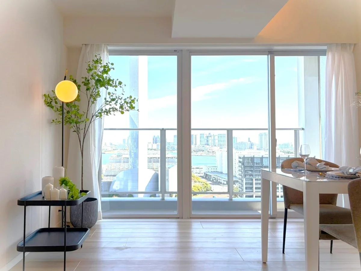 明るいリビングの部屋、窓から都市の景色が見える。テーブルにはワイングラスと食器が置かれている。左側に観葉植物とシンプルな飾りがある。