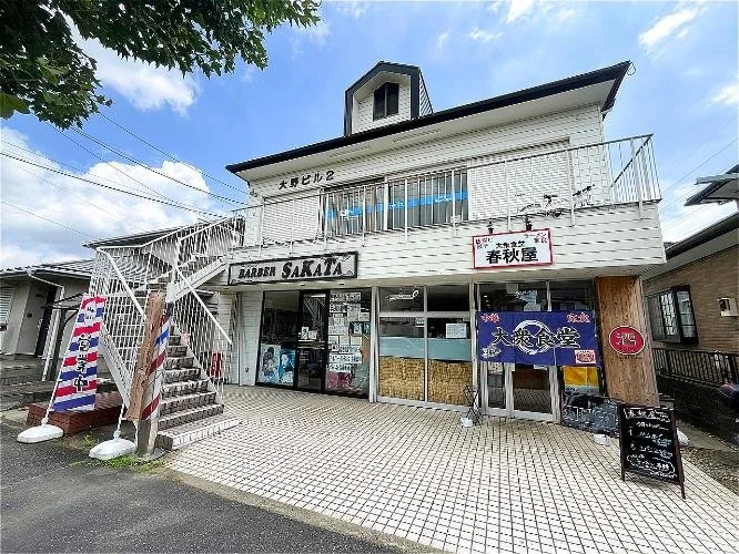 白い二階建ての建物で、看板に「BARBER SAKATA」と書かれています。階段の横にバーバーバッグがあります。屋上に小さな窓と塔がついています。