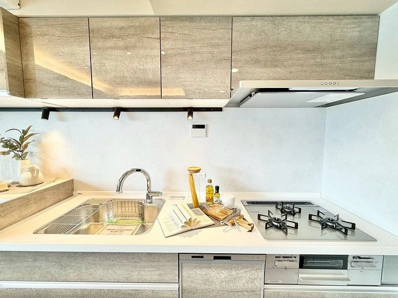 白いカウンターのキッチンで、シンクと三つのガスコンロがある。カウンターには調理用品と本が置かれている。上部に木目調の収納棚があり、左側には植物が置いてある。