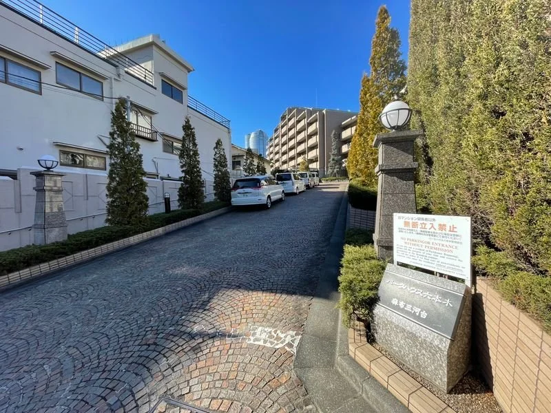 傾斜した石畳の坂道の右側に、フェンスと樹木が並び、左側には白い建物があります。坂道の先に複数の車が駐車されており、遠くにはビル群が見えます。