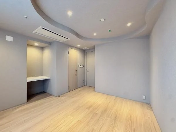 空のリビングルームで、木製のフローリングと白い壁、天井に間接照明とスポットライトがある。壁に小さな棚と天井にあるエアコンが見える。