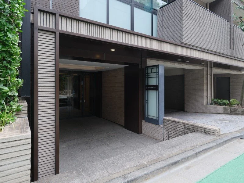 都会の建物のエントランスの入口。ガラスの扉と周囲に木材やコンクリートの壁。道路と歩道の一部も見える。