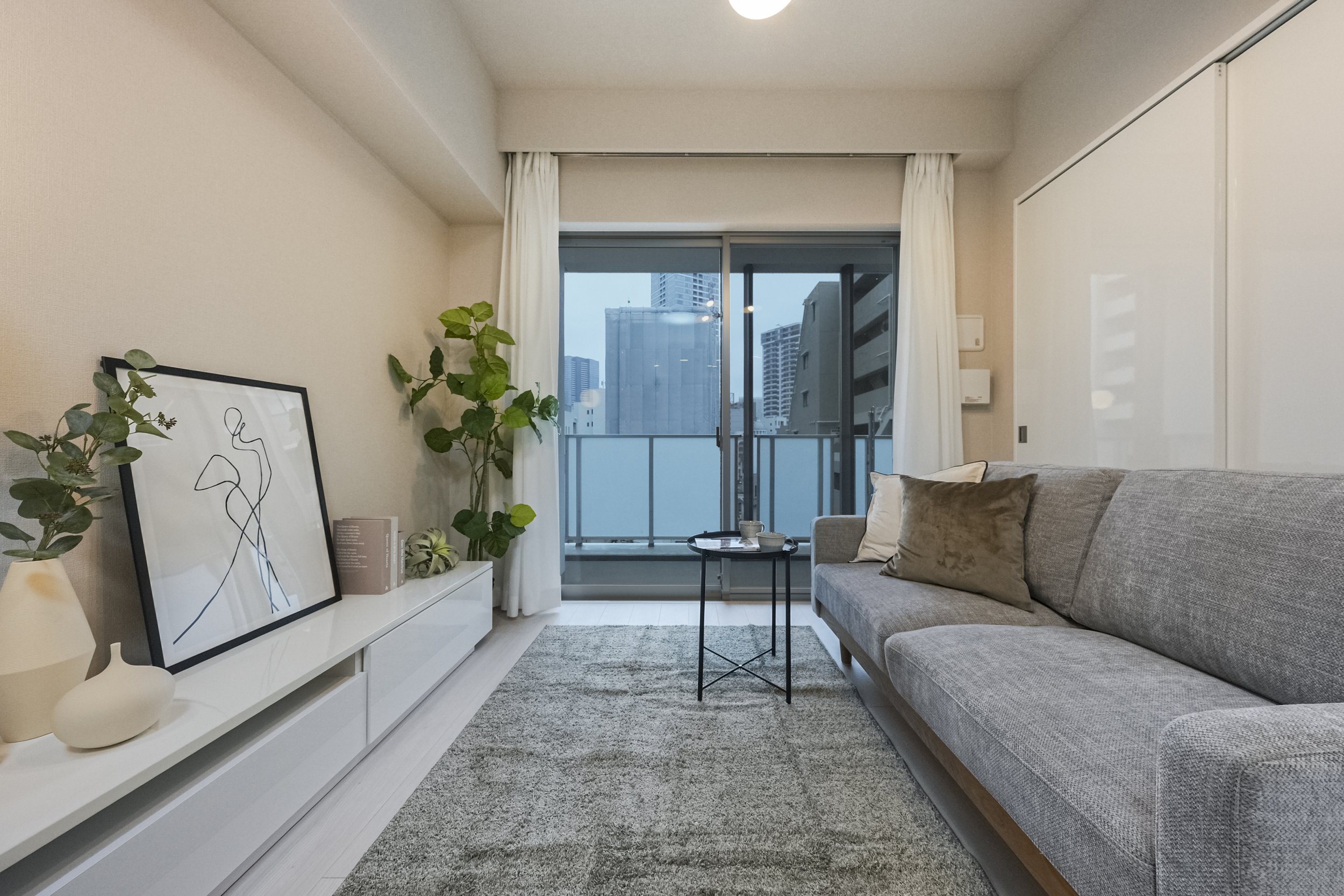 都市の高層ビルが見えるバルコニー付きリビングルームの内観。灰色のソファ、コーヒーテーブル、白いテレビ台と装飾品、観葉植物、カーテンがあり、自然光が入っている。