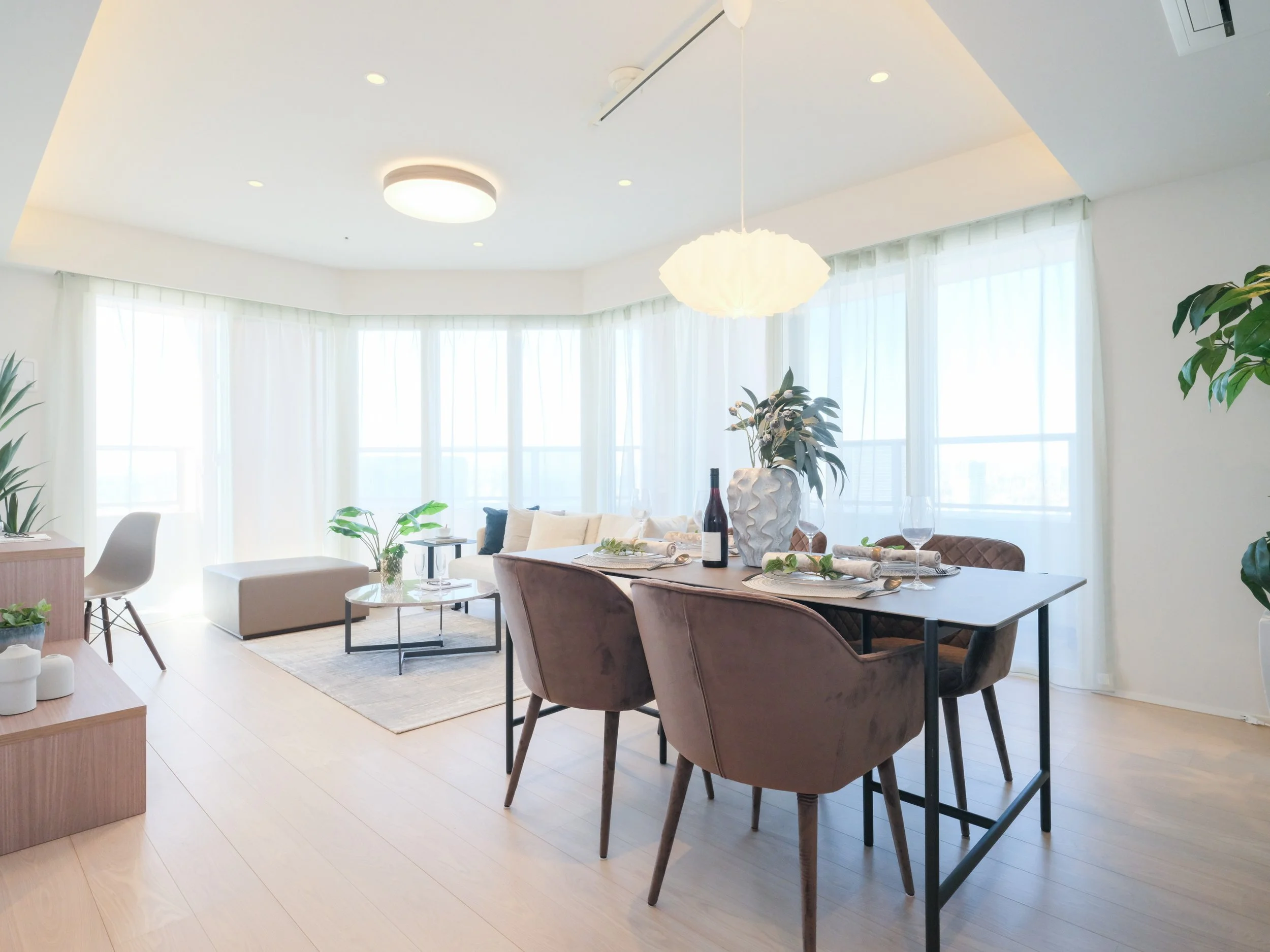明るいリビングとダイニングエリアの写真。大きな窓から自然光が入り、植物や家具が配置されている。ダイニングテーブルにはワインと食器がセットされている。