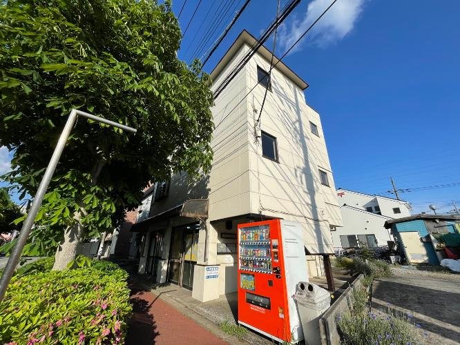 三階建ての白い建物と自動販売機、周囲に木と花があり、青い空と電線が背景に見える風景