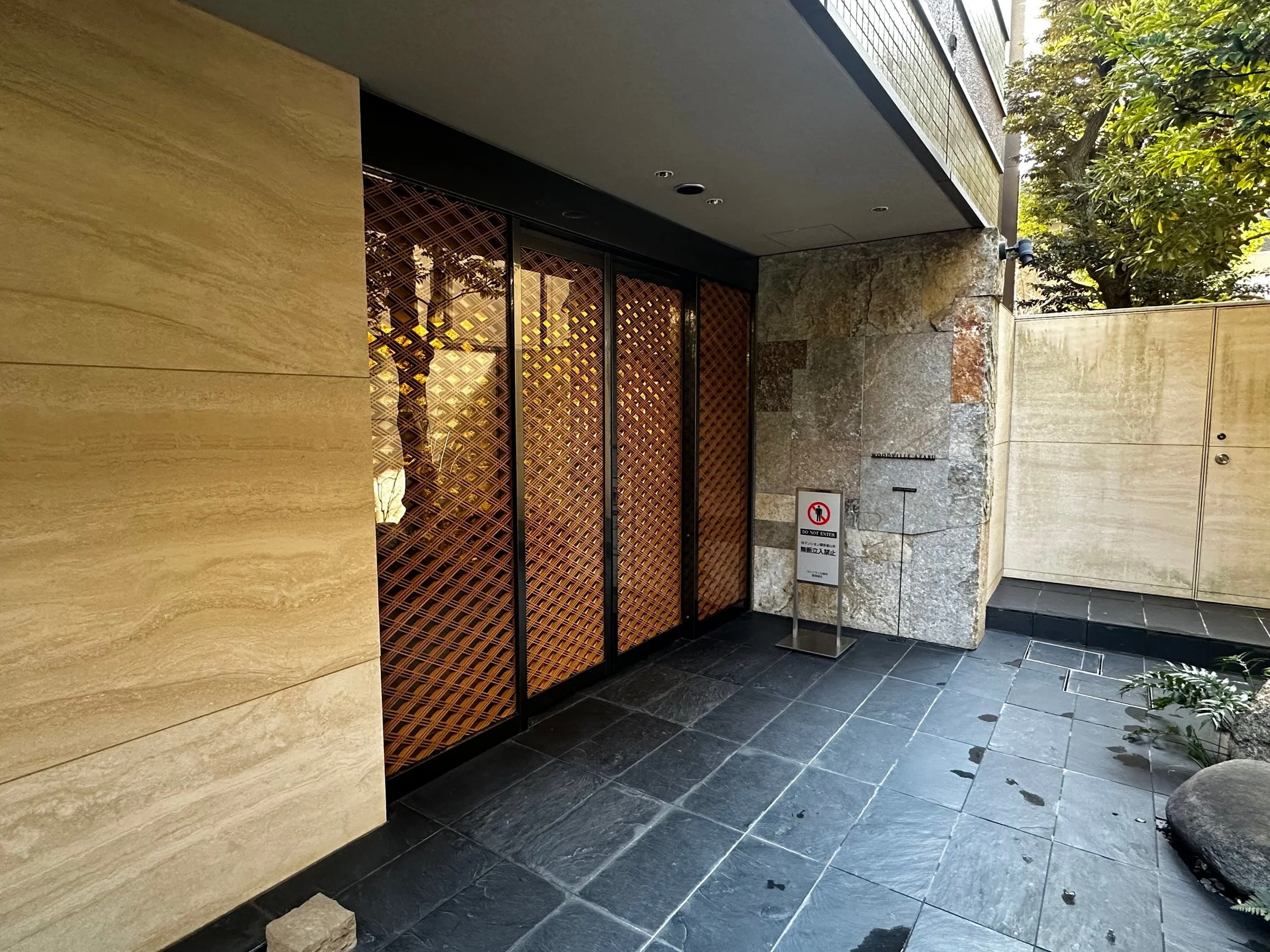 ビルの入り口に木製の格子模様の扉と「立ち入り禁止」の看板があり、黒い石の床と石壁、背後に緑豊かな木々が見える。