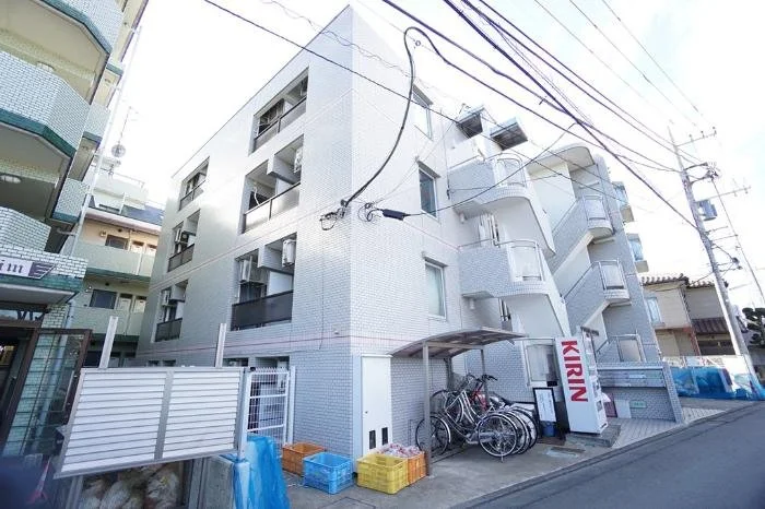 白い四階建てのアパートメントの外観。自転車置き場とバイク、プラスチックのコンテナが並ぶ。電線と電柱が上部に見える。
