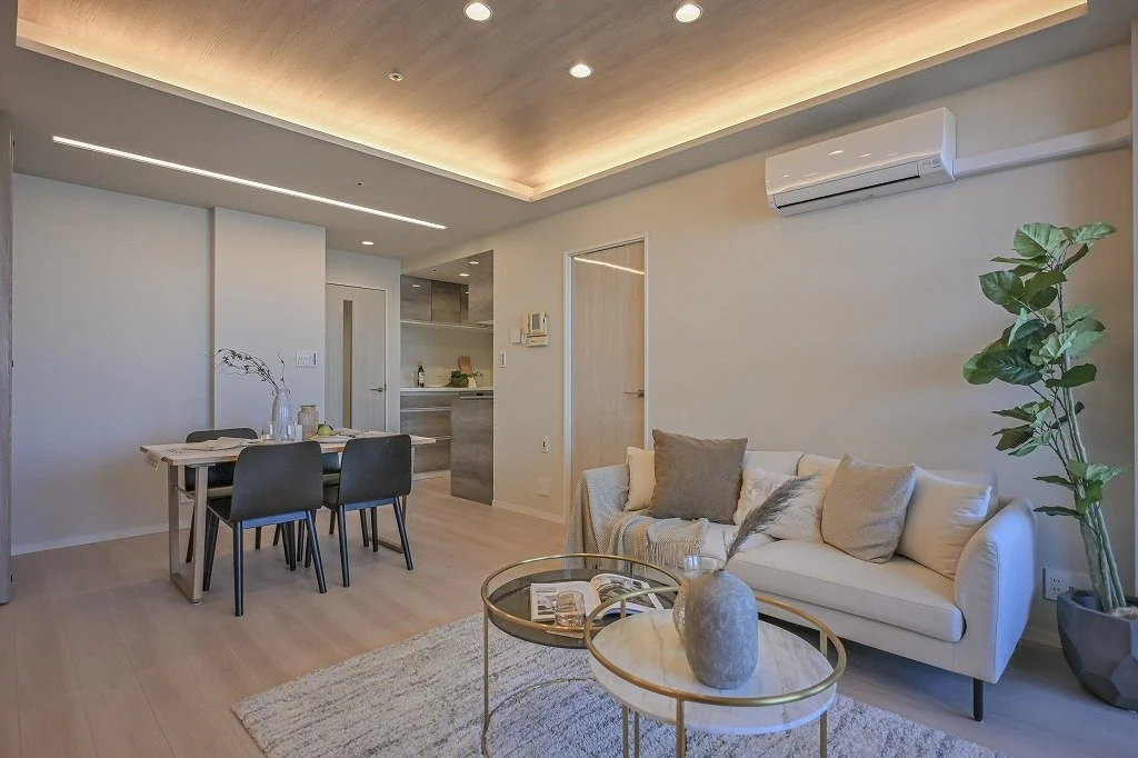 モダンなリビングルームとダイニングルームのインテリア。白いソファ、ラウンドテーブル、グリーンの観葉植物、木製のダイニングテーブルと黒い椅子、照明付きの天井とエアコン。