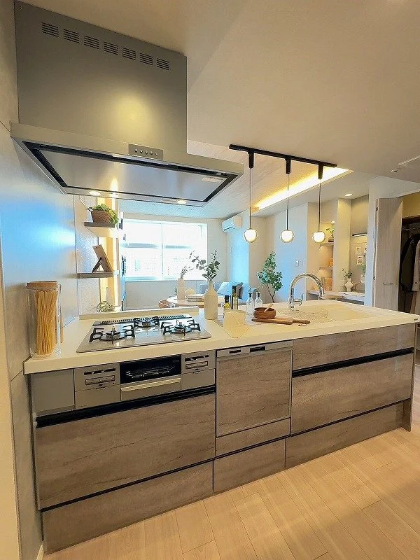 モダンなキッチンで、ガス炉、シンク、照明が設置されており、カウンターにはパスタや調理器具が置かれている。背面には窓と観葉植物があり、自然光が差し込む明るい空間。