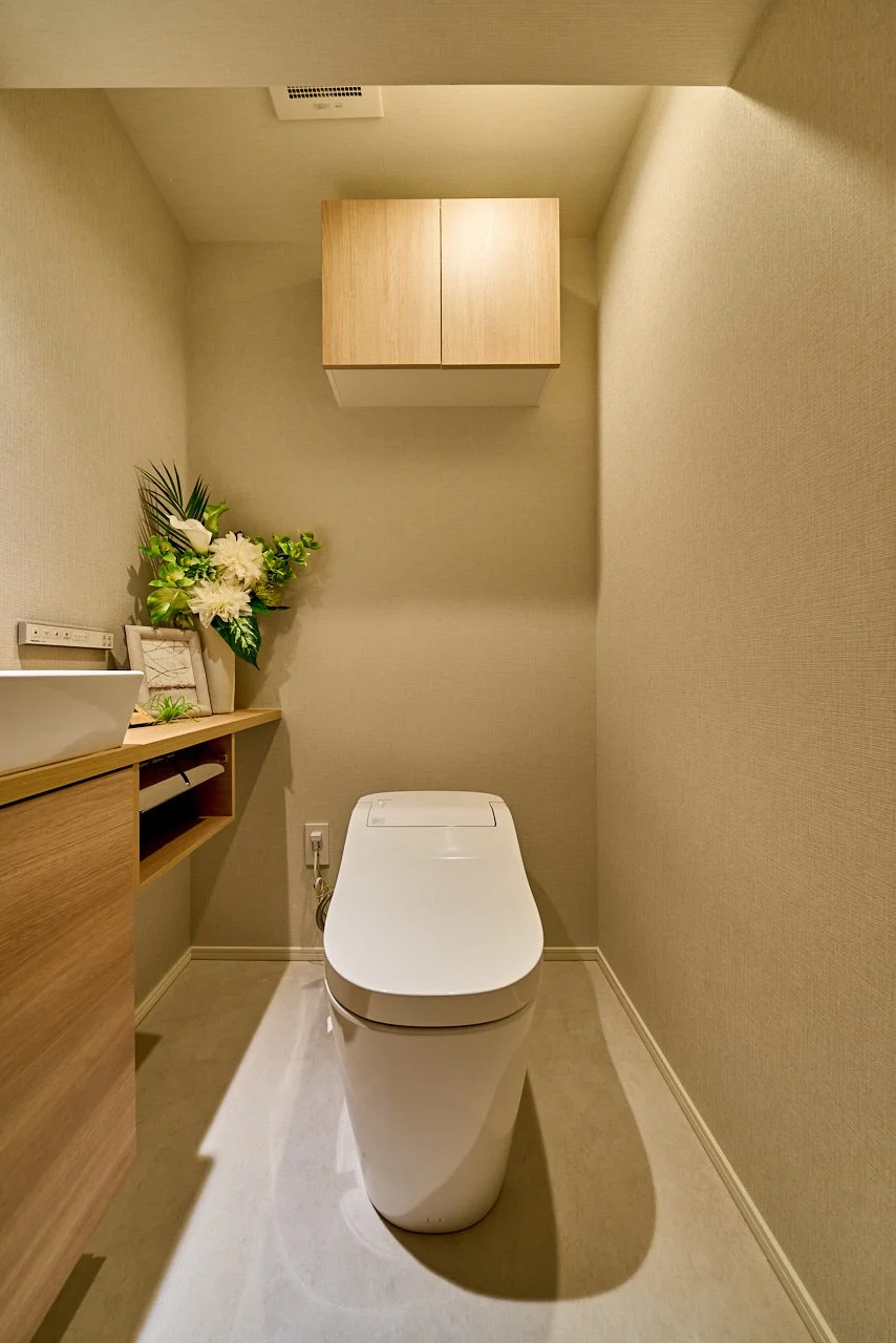 白い便座がある小さなトイレの部屋。左側には花の飾りと棚があり、天井に換気扇がついている。